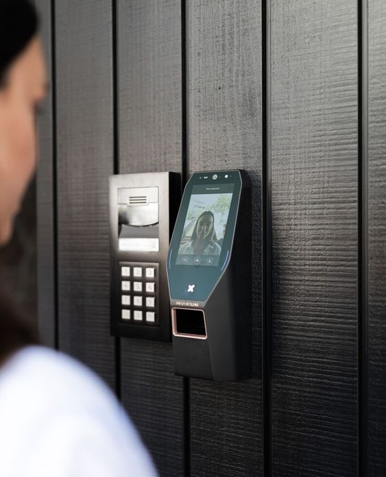 Woman using a modern facial recognition and keypad entry system on a black panel for secure access to a building.