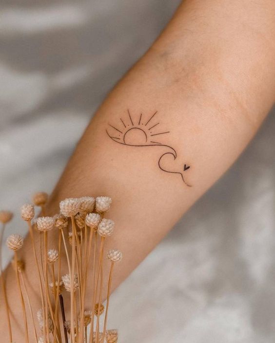 Minimalist tattoo of the sun, a wave, and a heart on an arm with dried flowers in the background. Simplicity and beauty.