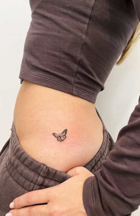 Close-up of a minimalist butterfly tattoo on a woman's right hip, wearing a brown cropped top and matching sweatpants.