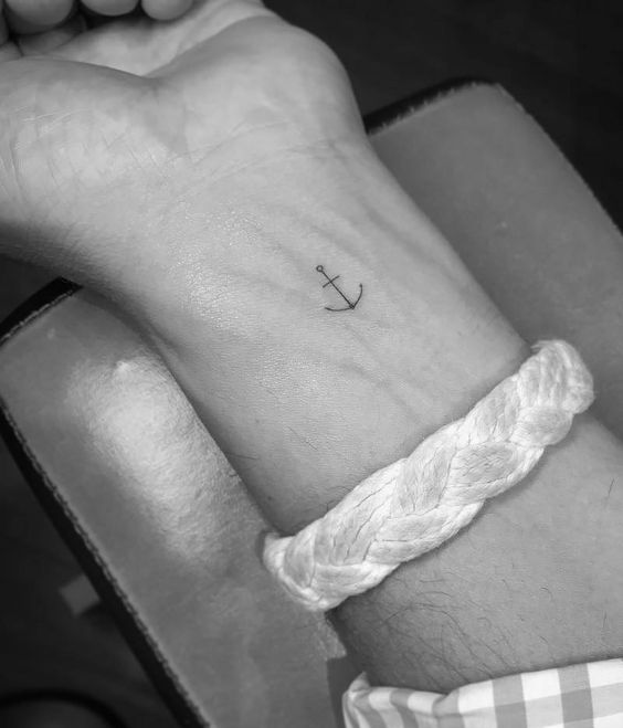 A small anchor tattoo on a wrist, accompanied by a braided white bracelet. Perfect for subtle nautical-themed body art.