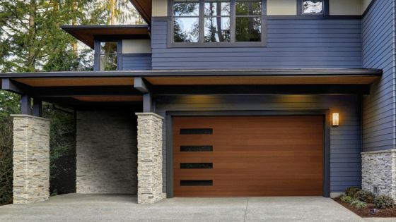 Modern house exterior with blue siding, stone accents, and a wooden garage door in a forested setting.