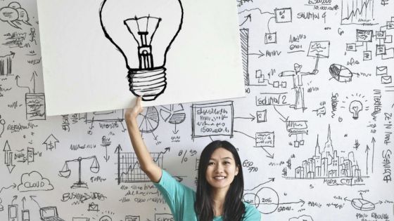 Woman holding a light bulb illustration in front of a whiteboard covered with brainstorming sketches and diagrams.