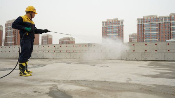 Worker in protective gear using a pressure washer to clean a concrete surface on a rooftop with city buildings in the background.