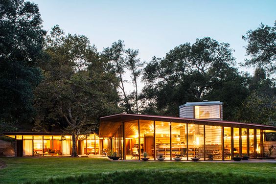 Modern glass house with warm interior lighting nestled in a forested area at dusk, showcasing contemporary architecture.