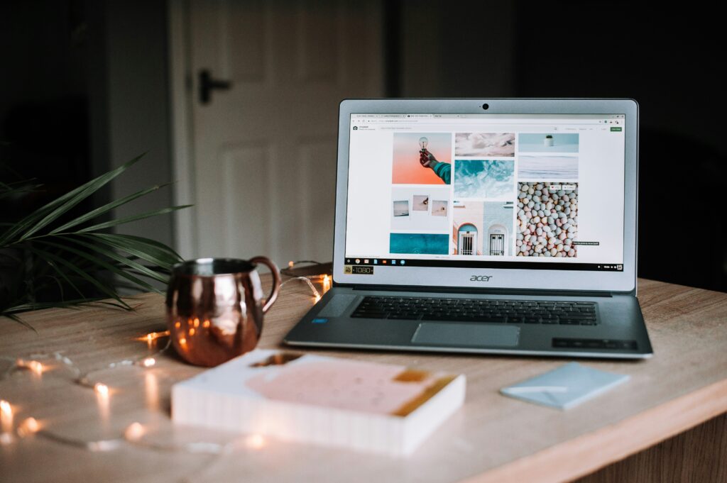 Acer laptop on wooden desk, open to a photography blog, with notebook, copper mug, string lights, and plant nearby.