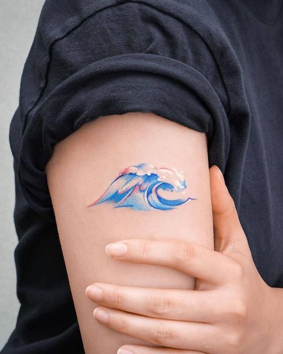 Close-up of a colorful ocean wave tattoo on an upper arm, designed in blue and pink hues, with a rolled-up sleeve.