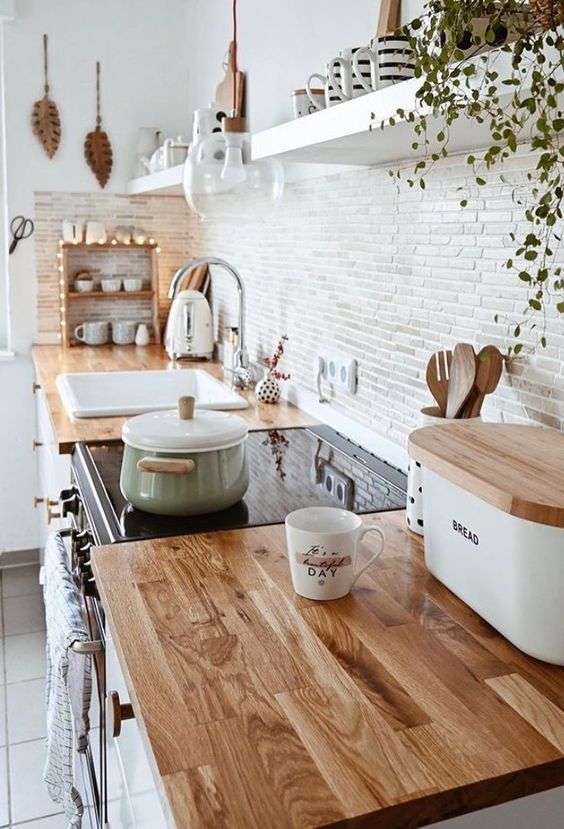 Modern kitchen with wooden countertops, white cabinets, and shelves, featuring cookware, a bread box, and hanging plants.