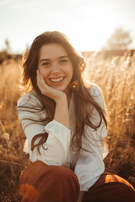 Woman smiling in golden field at sunset, wearing white blouse and brown pants, enjoying nature and warm sunlight.