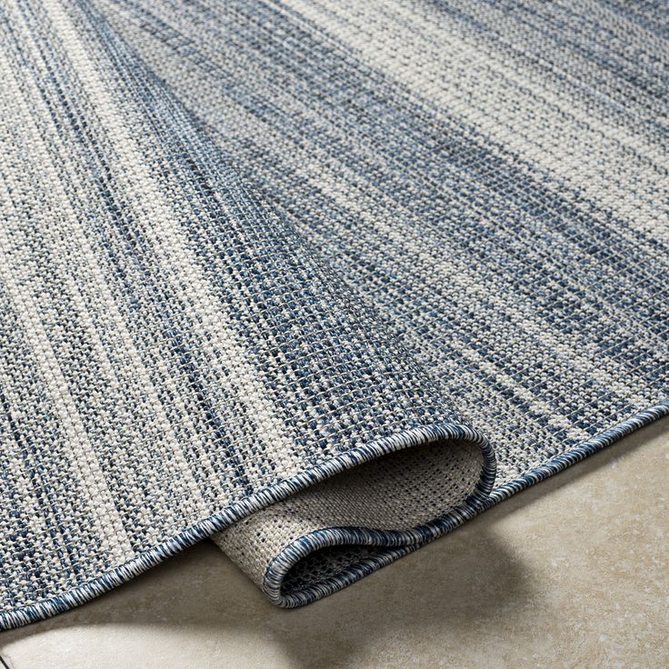 Close-up of a blue and grey woven floor rug with a rolled edge on a beige tile surface.