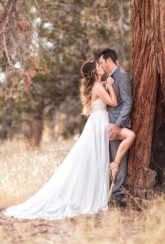 Bride in flowing white dress kissing groom in grey suit by a tree in a serene outdoor setting.