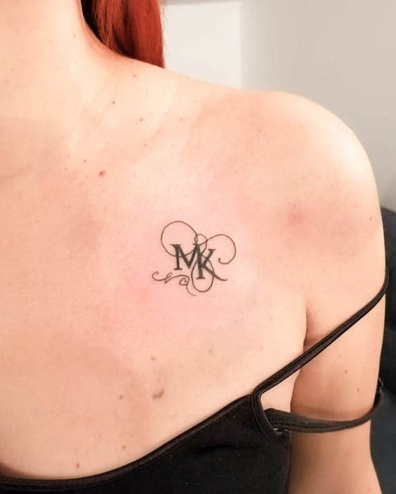 Close-up of an intricate MK monogram tattooed on a woman's upper chest, near her shoulder, wearing a black spaghetti strap top.