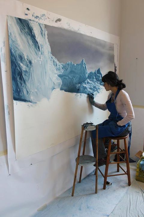 Artist painting a detailed glacier on a large canvas in a studio, wearing an apron and seated on a stool.