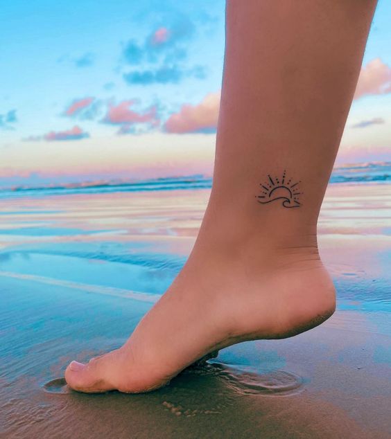 Bare foot with sun and wave tattoo on ankle at beach during sunset, reflecting sky and clouds in water.