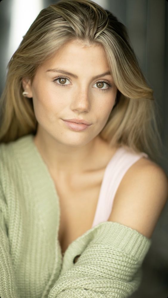 Young woman with long blonde hair, wearing a light green sweater and a tank top, looking directly at the camera.