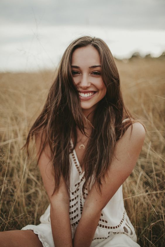 Woman smiling in a white dress, sitting in a golden field of tall grass on a cloudy day, capturing a joyful outdoor moment.