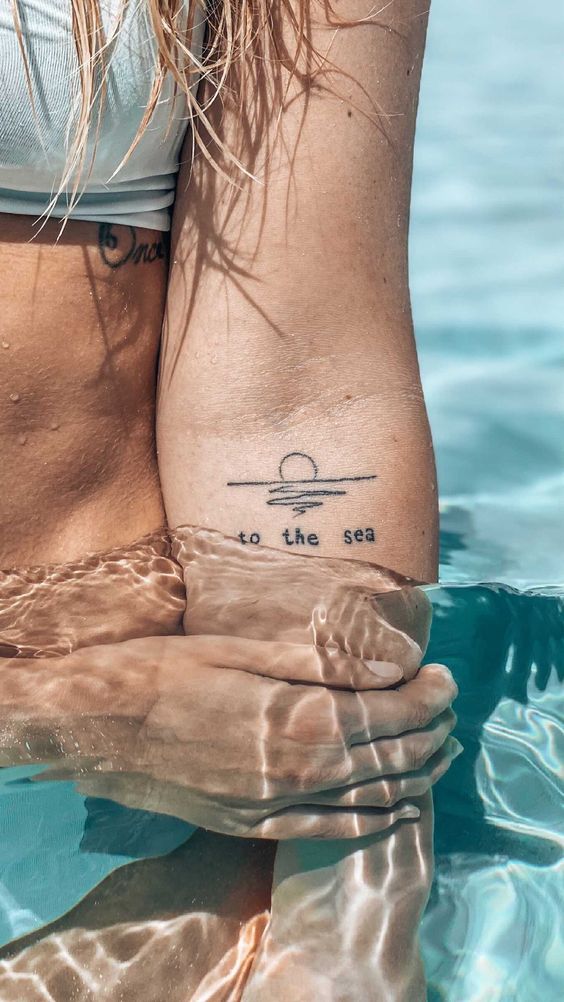 Close-up of a person in a pool with a to the sea tattoo on their arm, reflecting sunlight on rippling water.