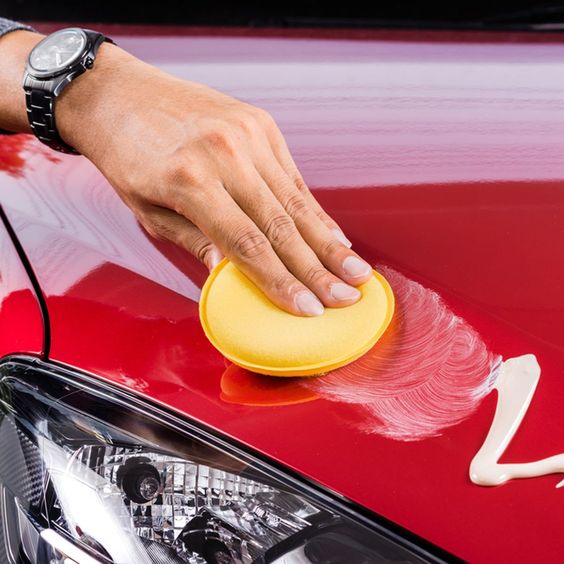 Hand polishing red car's hood with yellow sponge and cream wax near headlight.