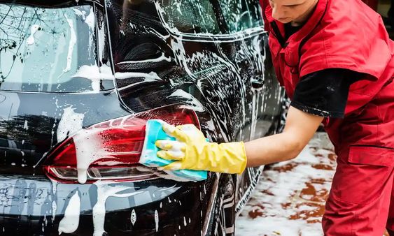 Person washing a black car with a sponge and soapy water, wearing yellow gloves and red uniform.