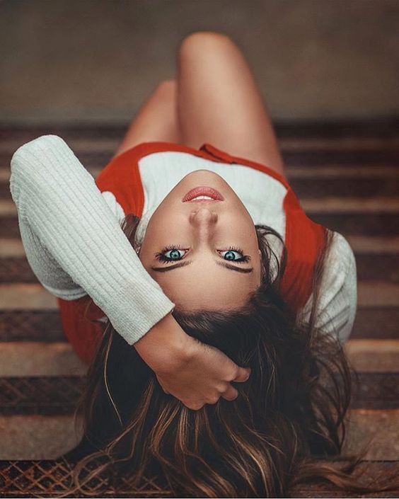 Woman with blue eyes lies on stairs, wearing a white sweater and red vest, hand in hair. Close-up, looking at camera.