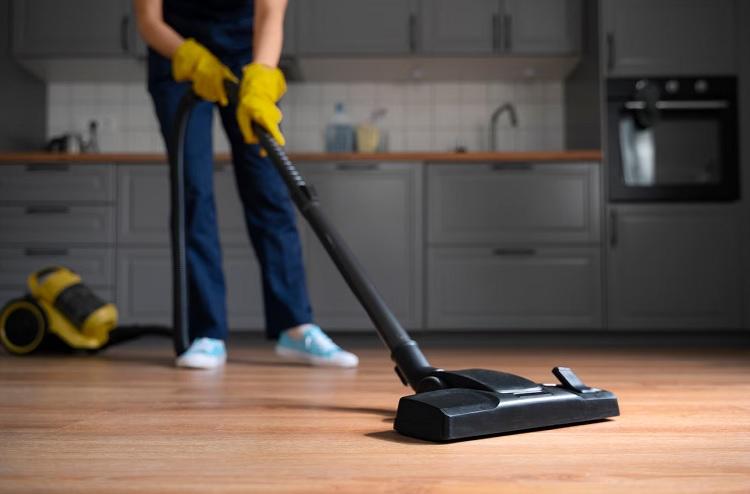 Person vacuuming a kitchen floor with a black vacuum cleaner, wearing blue pants and yellow gloves. Modern kitchen in background.