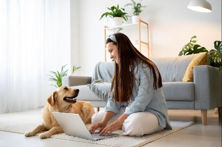 Woman working on a laptop while sitting on the floor with her dog in a cozy living room with green plants. Working from home.