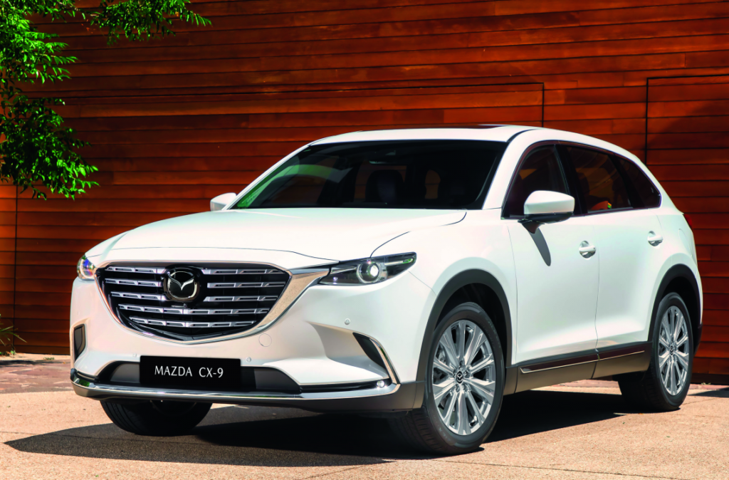 White Mazda CX-9 SUV parked in front of wooden wall with green foliage, showcasing modern design and sleek appearance.