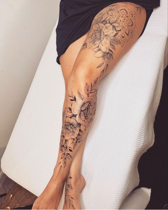 Detailed floral leg tattoo with mandala and leaf designs on thigh and calf, showcased on a woman lying on a white surface.