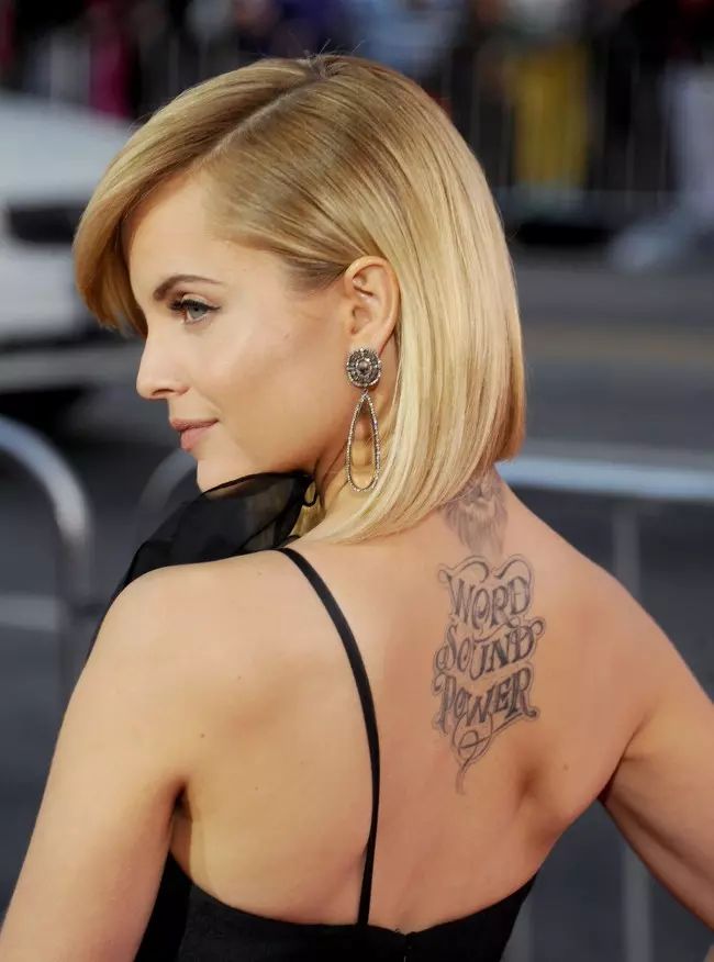 Blonde woman with short hair shows back tattoo saying Word Sound Power while wearing black dress and dangling earrings.