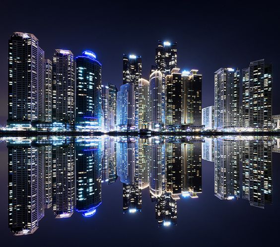 Modern city skyline reflecting in calm water at night, with illuminated skyscrapers creating a stunning mirrored effect.