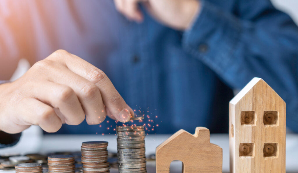 Hand stacking coins next to wooden house models, symbolizing real estate investment, savings, and financial growth.