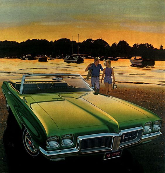 Green classic convertible at sunset by the lake with a couple walking in beachwear, boats docked in the background.