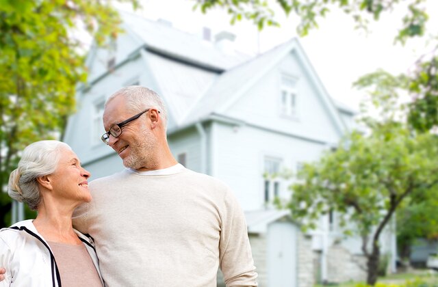 Elderly couple smiling at each other in front of their house surrounded by trees, depicting homeownership and happiness.