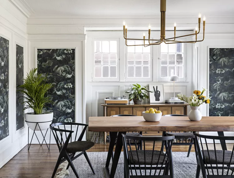 Modern dining room with wooden table, black chairs, chandelier, plants, floral wallpaper, and bright natural light.