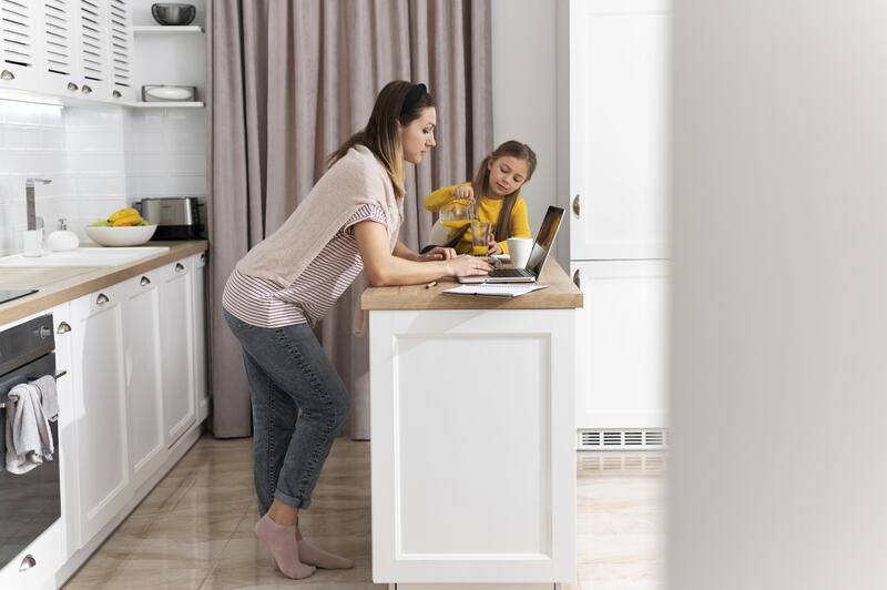 A woman working on a laptop in a modern kitchen while a child pours milk into a cup beside her.