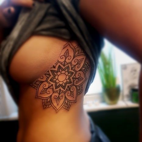 Mandala rib tattoo on a woman's side in a flower design, with intricate details. Upper body close-up, casual setting.