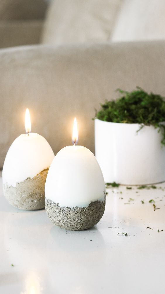 Minimalist egg-shaped candles with concrete base lit on a table, complemented by a small potted plant in the background.