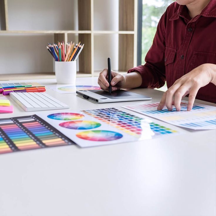 Designer using a graphics tablet and color swatches at a desk filled with pencils, keyboard, and color charts.