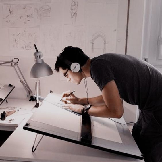 Architect drawing on drafting table, wearing headphones, focused on work in a creative studio with sketches on the wall.