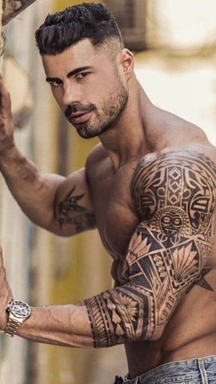 Muscular man with intricate arm tattoos posing shirtless outdoors.