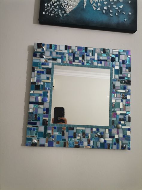 Square mirror with a colorful mosaic frame made of blue and white tiles, reflecting a person taking a photo with a phone.