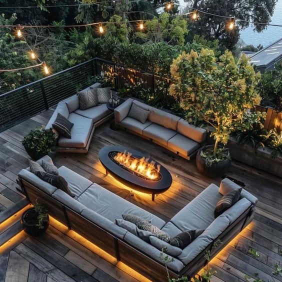 Modern outdoor patio with sectional sofas, fire pit, string lights, and lush greenery overlooking a scenic view at dusk.