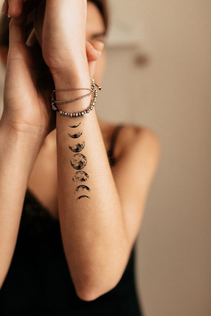 Arm with moon phase tattoo and layered bracelets on wrist, against a neutral background.