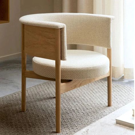 Modern beige upholstered chair with wooden frame, placed on a textured rug in a bright room with natural light filtering through curtains.