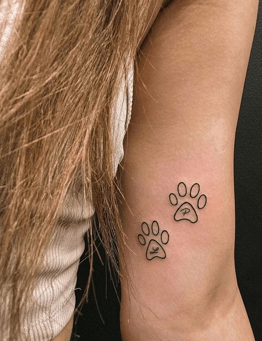 Two minimalistic paw print tattoos on a woman's upper arm, featuring initials L and P inside the prints.