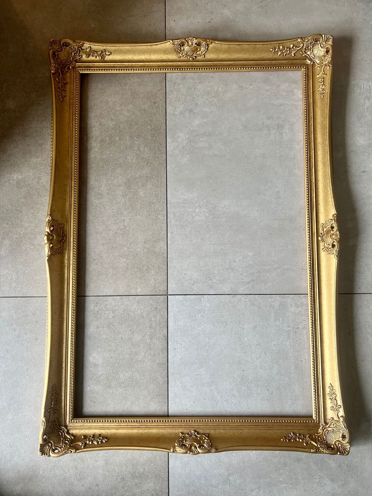 Vintage ornate gold picture frame with intricate detailing, displayed on a gray tile floor background.