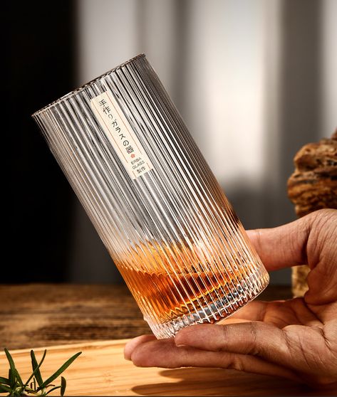 Hand holds an elegant ribbed glass tumbler with amber liquid inside, on a wooden table, minimalist aesthetic.