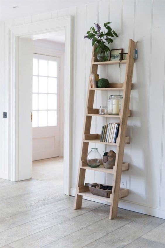 Minimalist wooden ladder shelf with books, plants, and decor against white paneled wall in bright, modern home interior.