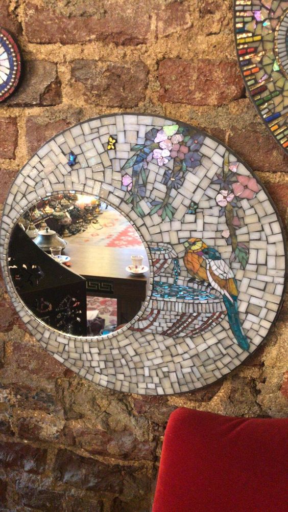 Round mosaic mirror with floral and bird design hangs on exposed brick wall above red chair in cozy room.