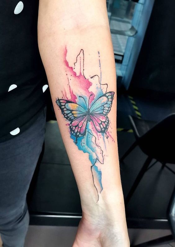 Colorful butterfly tattoo with watercolor background on a person's forearm, blending vibrant blue, pink, and purple hues.