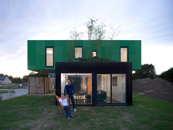 Modern two-story green and black house with couple outside sitting and standing on lawn. Contemporary architecture with large windows.